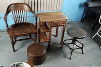 Set showing wooden courtroom chair, small wooden desk, brown vinyl footrest, and industrial metal stool on concrete floor.