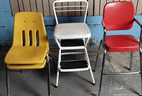 Two yellow plastic chairs, white metal Cosco step stool, and small vintage red chair placed side by side on concrete floor with blue wall background.