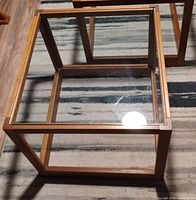 Two cube-shaped end tables with wooden frames and clear glass inserts on top and bottom surfaces shown from above and side angles on a rug.