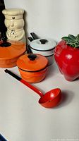 Image showing the full assortment of cookware and ceramic canisters including two orange enamel pots, white pot, orange ladle, ceramic chef canister, and red apple canister.