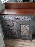 Front view showing the wooden top, three drawers decorated with floral pattern, and one cupboard door also with floral pattern. Metal handles and knob visible. Cabinet body is painted gray with natural wood top.