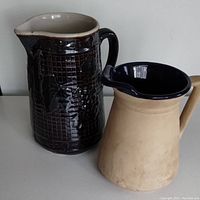 Two rustic ceramic pitchers side by side showing color and design details: dark brown textured pitcher with white interior, and beige-dark blue pitcher.