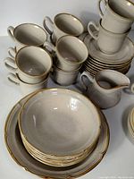 Photo showing stacked coffee cups with handles, matching round saucers, rimmed cereal bowls, and a small creamer pitcher all in beige stoneware with brown rims.