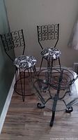 Photo showing two black wrought iron swivel stools with decorative scrollwork backs and cushioned black and white patterned seats next to glass top wrought iron table.