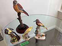 View of five bird figurines displayed on glass table; includes tall resin bird on stand and four smaller porcelain/china birds.
