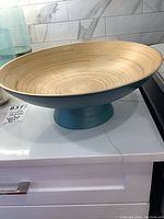 Painted bamboo footed bowl with teal blue exterior and natural bamboo interior on a white counter; visible crack on the counter around the base of the bowl.