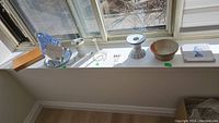 Seven items lined on a window sill: wood box, blue and white pottery, two white candle holders, pottery bowl, toothbrush holder, hummingbird feeder.