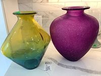 Two large glass decorative vases side by side on a white countertop, one green-yellow multicolored and one purple.