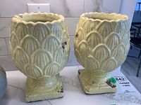 Pair of yellowish ceramic vases with scalloped texture and square pedestal base shown together from a slightly front angle.