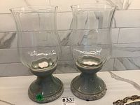 Two Bombay hurricane lamps displayed side by side showing ceramic bases with blue-green glaze and clear glass chimneys.
