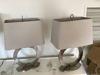 Full view of two table lamps, showing brushed metal circular bases with beige rectangular fabric shades placed on a white surface.