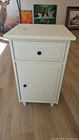 Front view of white Ikea night table showing drawer and lower cabinet door, both with black knobs, standing on short rounded feet.