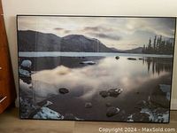 Framed photo print propped against wall showing serene mountain lake scene with rocks and distant trees.