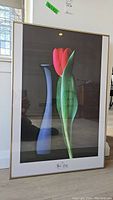 Framed floral print showing abstract red and green tulip against dark background with light wooden frame.