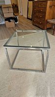 Square glass top side table with chrome metal legs, showing clear glass surface with chip on one corner, placed on carpeted floor with wooden furniture and boxes in background.