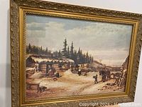 Close-up of framed print showing the winter cabin scene with people and horses, ornate gold frame visible