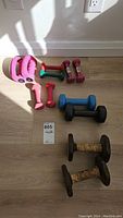 Photo shows multiple dumbbells on a wood floor against a white wall with outlet, including pink, green, purple neoprene dumbbells, blue and black rubber dumbbells, and rusted metal dumbbells with wrapped grips.