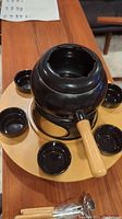 Black fondue pot with wooden handle on a wooden Lazy Susan tray with six small black bowls arranged around it.