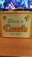 Front view of the Orange Crush lunch box with embossed text and floral decoration on lid.