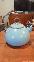 Front view of a light blue tea kettle with stainless steel handle and attached whistle on its spout.