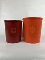 Front view of the two large cylindrical Tupperware storage containers side by side, one harvest orange with swirl emblem, one red tulip with floral emblem.