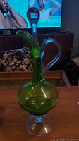 Angle view showing the elegant green glass decanter with clear glass swan handle and stopper, standing on wooden table.