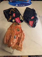 Three baseball gloves on a surface: Two black Rawlings gloves and one brown Rawlings glove.