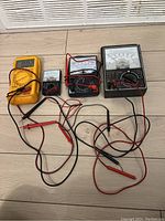 Four electrical test meters including a yellow digital multimeter and three analog multimeters with probes and leads.