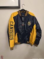 Full view of Canisius bomber jacket in navy blue and yellow hanging on black hanger against beige wall.