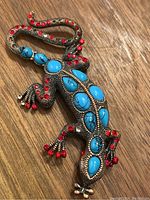 Top view of the lizard brooch showing turquoise-blue oval stones and red round stones on its body and tail, placed on a wooden surface.