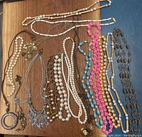 Top view of 12 assorted necklaces spread out on wood surface showing various beads, chains, pendants and colors including white, pink, blue, brown and gold tones