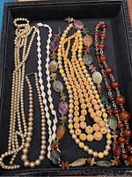 Photo showing seven assorted beaded necklaces arranged in a black box. Necklaces vary in bead size, color, and material.