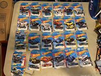 Top down view of 26 packaged Hot Wheels die-cast cars arranged in rows on a table.