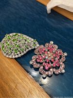 Two brooches side by side, one leaf-shaped with green and clear rhinestones, one flower-shaped with pink and clear rhinestones