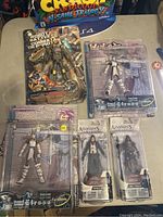 Lot of 5 action figures visible on table with original packaging. Includes Robotech, Warrior, and Assassins Creed themed figures.