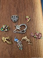 Lot of 8 brooches displayed on wood surface, showing variety of designs including floral, animals, shoe and abstract knot shapes.