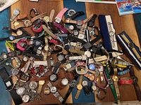 Top-down view of around 50 assorted watches piled together on a wooden surface showing variety of styles, bands and watch faces.