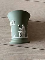 Side view of small matte green Wedgwood Jasperware vase showing white relief figures of a woman with staff and putti beside trees and flowers