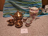 Full lot with gold Royal Winton teapot, sugar bowl, creamer, tall Bavaria floral teapot, and three floral cups on carpeted floor.