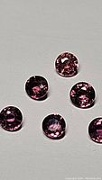 Six round, calibrated 3mm sapphires in varying fancy colors including deep pink, purple-pink and orange hues shown on a light gray surface.