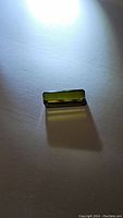 Bright green rectangular octagon cut tourmaline gemstone placed on white surface, showing size and color under direct light.