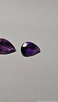 Two pear cut deep purple amethyst gemstones placed side by side on a white surface, showing color and faceting