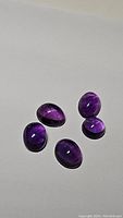 Five deep purple amethyst cabochon gemstones arranged on white surface, showing polished oval shapes and natural inclusions.