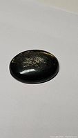 Oval cabochon cut gold sheen obsidian gemstone with visible gold sheen flash on a polished black surface.