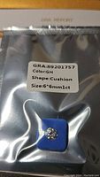 Packaged 1 ct cushion cut moissanite gemstone in sealed anti-tamper pouch showing GRA certification label with color GH, shape Cushion, size 6x6 mm.