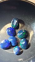 Seven faceted jet black Ethiopian opal gemstones shown together under light, displaying intense multicolor flashes and variations in size and color pattern.