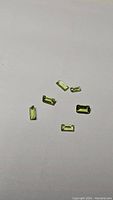 Six apple green baguette cut natural peridot gemstones displayed on a white surface, showing shape and color.
