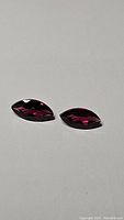 Pair of marquis cut pink purple Umbalite garnet gemstones placed on a grey surface with light reflections showing facets clearly.