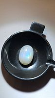 Oval translucent white moonstone with blue rainbow flash placed in a black circular holder, showcasing the stone's iridescent qualities.