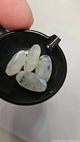 Four oval cut rainbow moonstones displayed in a small scoop, showing blue rainbow flashes and milky translucent bodies with natural inclusions.